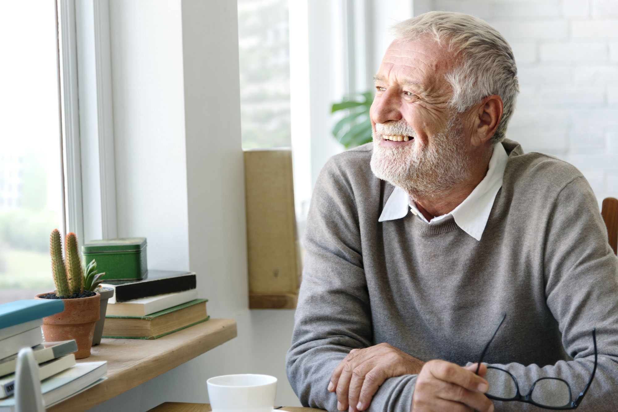 A smiling senior man looks out the window while enjoying coffee.